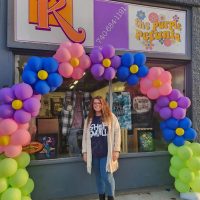 Purple Petunia owner Katy standing in front of the store with arc of balloons arranged like flowers.