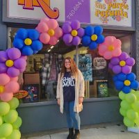 Purple Petunia owner Katy standing in front of the store with arc of balloons arranged like flowers.