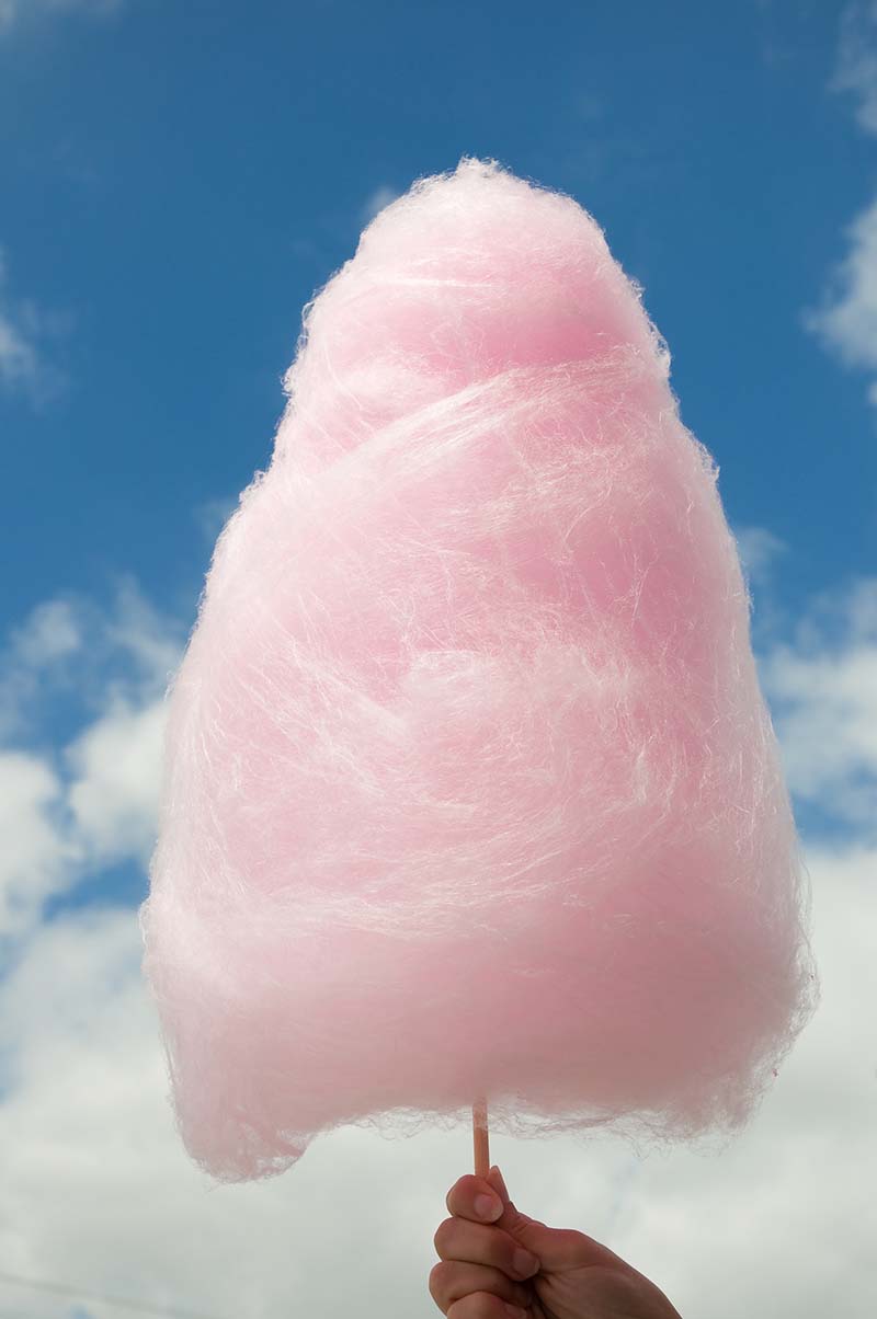 Pink mound of cotton candy on stick behind held by a hand with blue sky background.