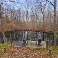 Two chairs sitting on deck at edge of small pond among tall trees at Honeybee Hideaway in Logan, Monday Creek, southern Perry County, Ohio