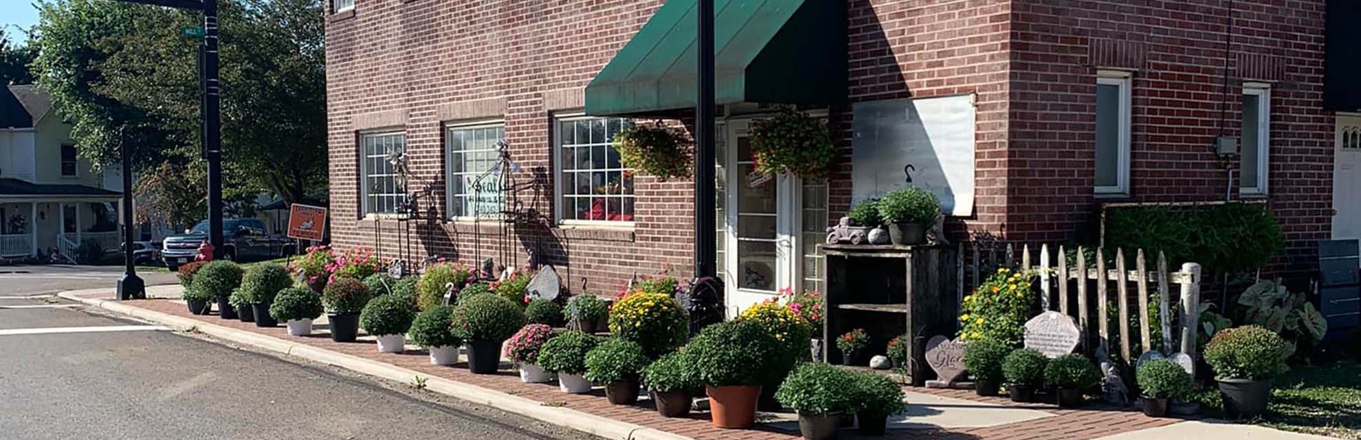 Red brick storefront with green awning entrance and a variety of large plants lining the sidewalk at Seals Flowers & Gifts in New Lexington, Perry County, Ohio.