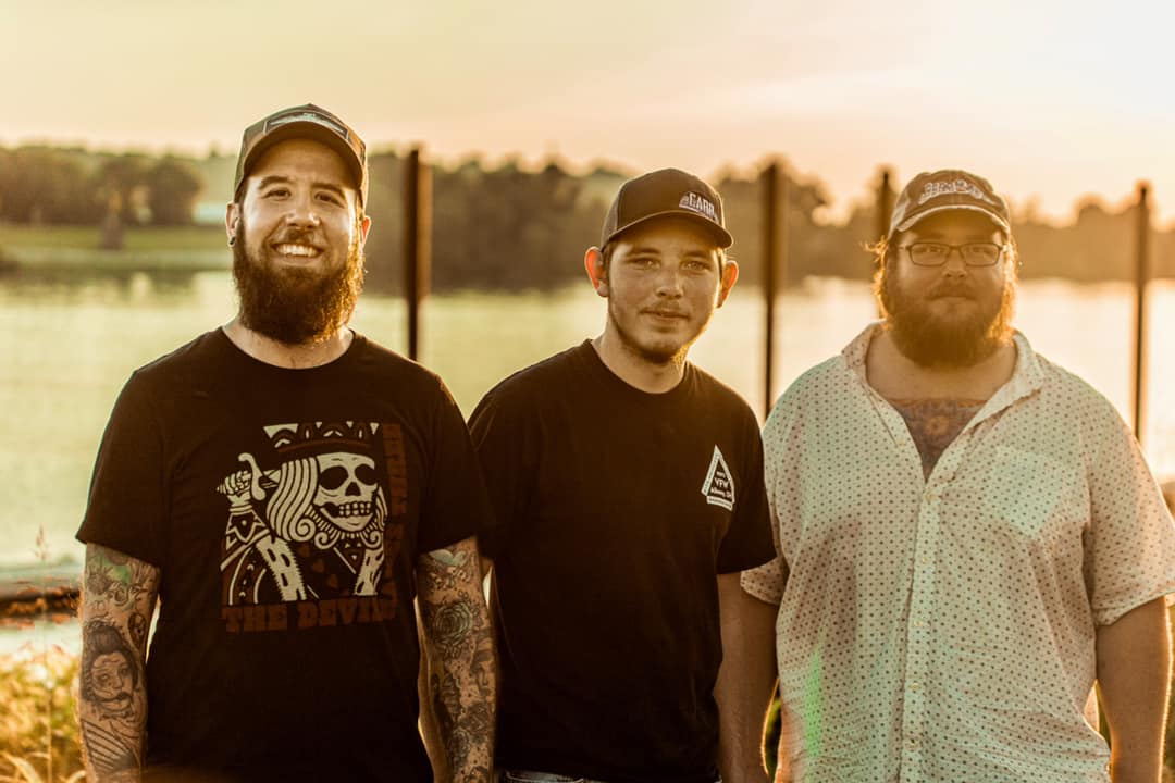 Three men, the Coal Cave Hollow Boys, standing together in t-shirts at sunset